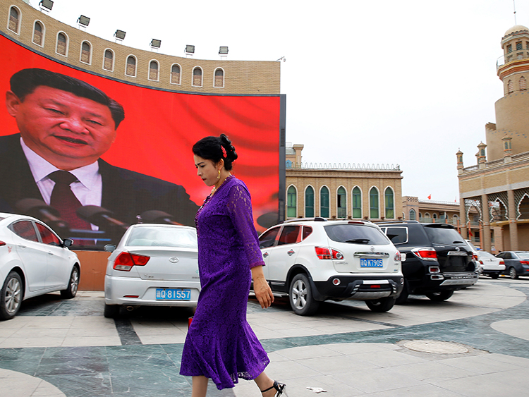 中国国家主席・習近平を映す大画面の前を歩くウイグル族の女性=2018年9月、中国新疆ウイグル自治区カシュガル、ロイター