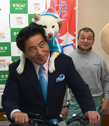自転車に乗る片岡聡一市長の背に乗る太郎と、飼い主の松本光正さん(右)=岡山県総社市役所